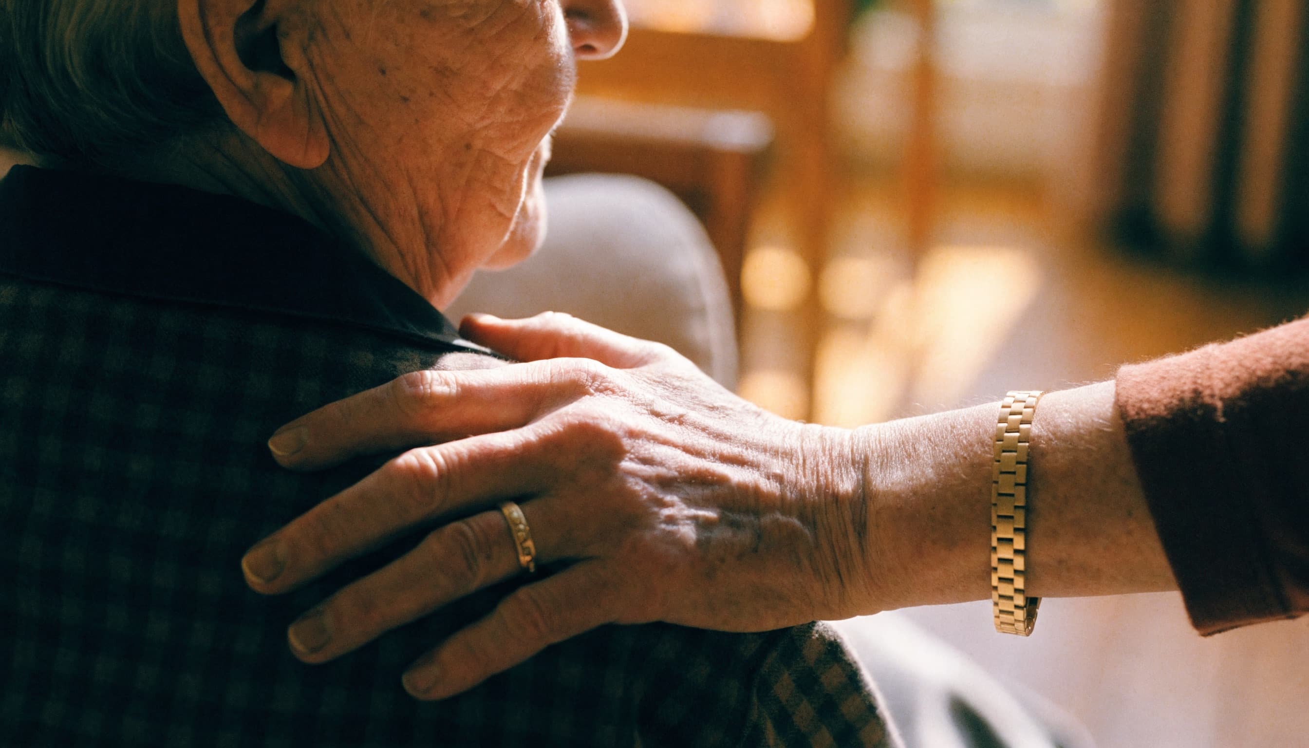 A carer's hand resting reassuringly on an elderly person's shoulder in a softly lit home