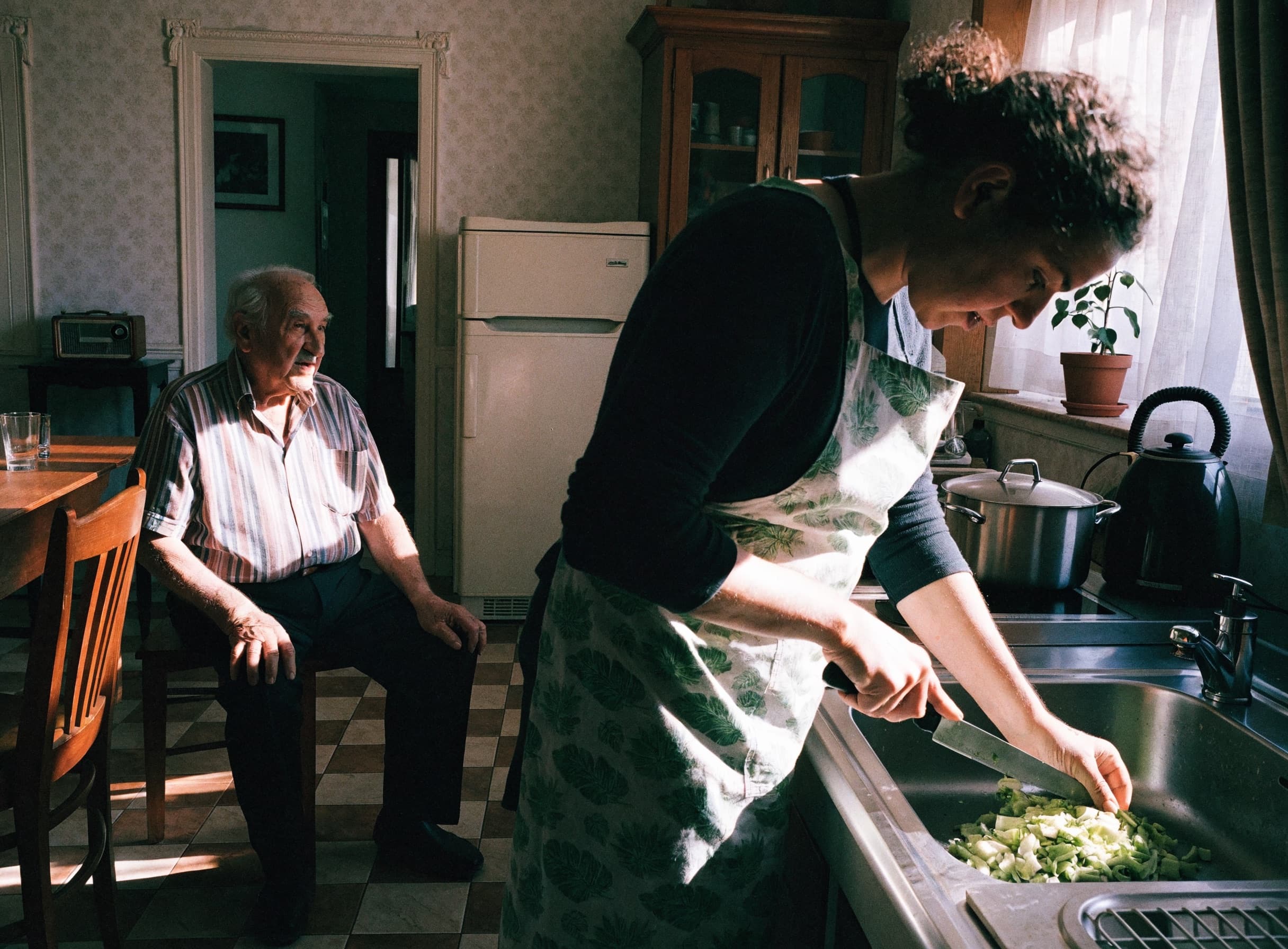 A home helper preparing a fresh meal in a sunlit kitchen while an elderly man chats nearby