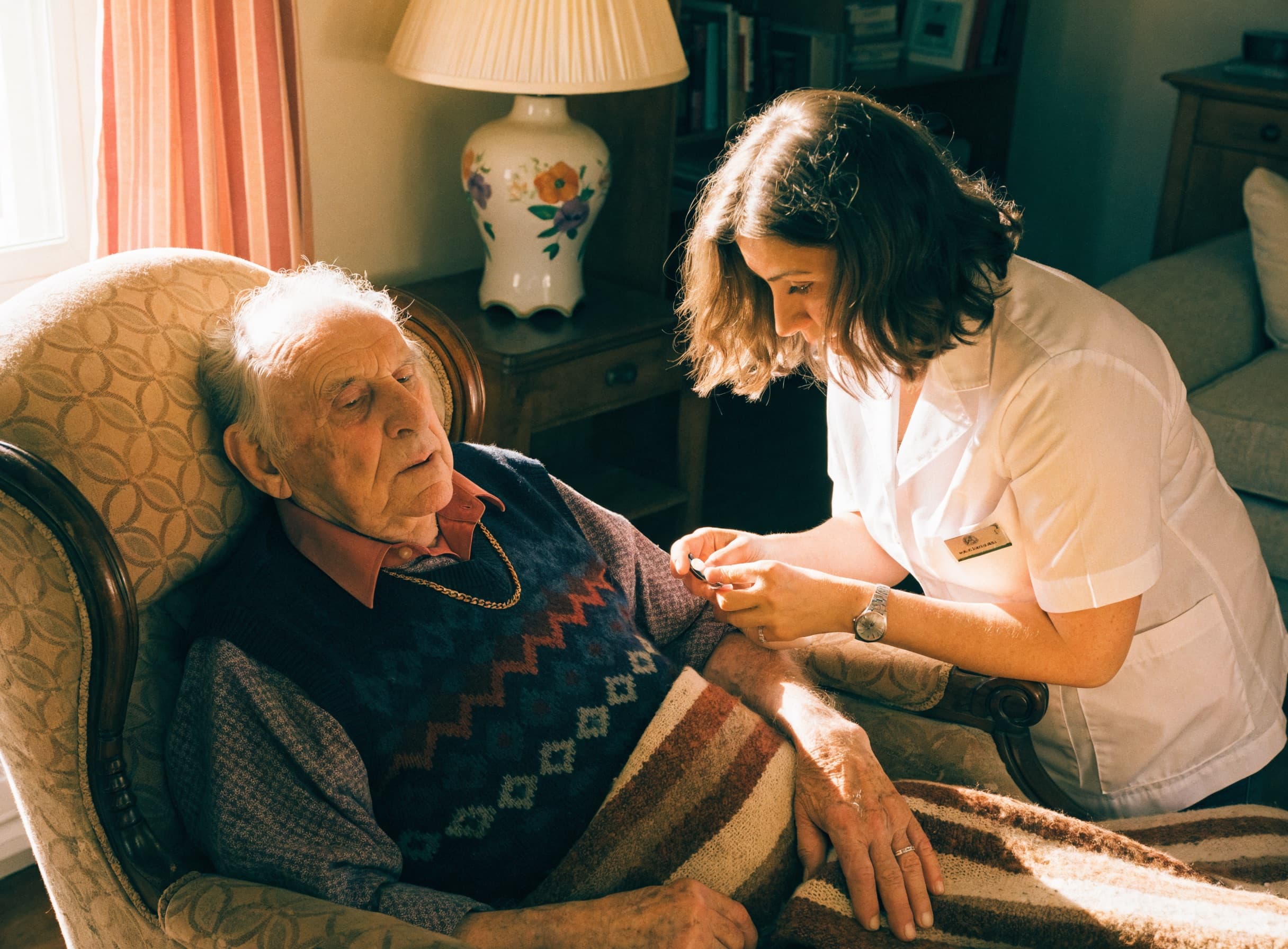 A gentle home nurse checking on an elderly man in a comfortable sunlit living room