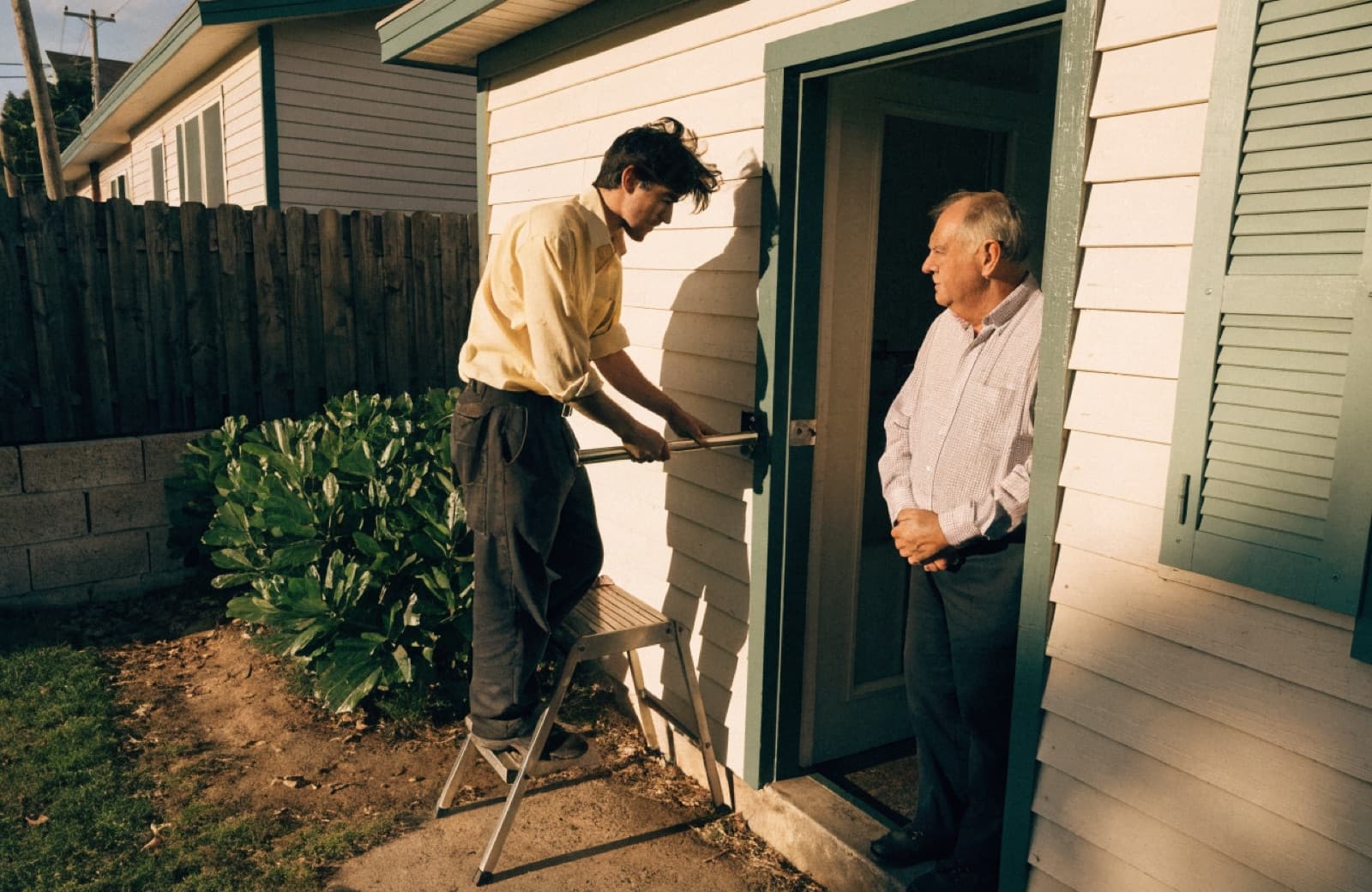 A handyman fitting a safety grab rail beside a back doorstep while an older man watches from the doorway
